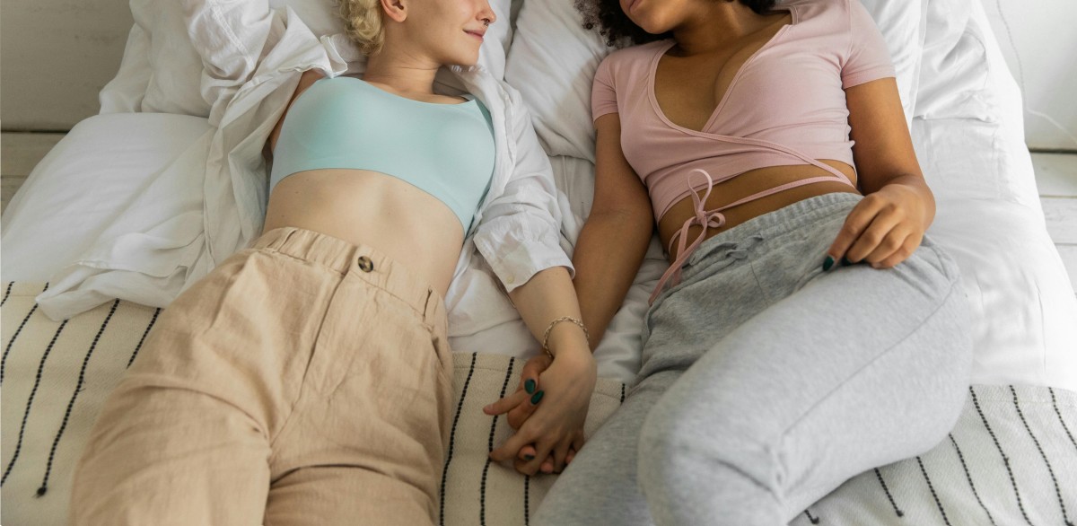 Two women laying in bed holding hands, Photo Credit: Pexels